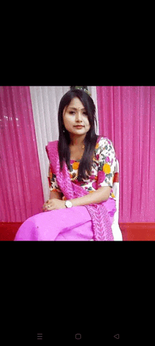 a woman in a purple saree is sitting in a chair in front of a pink curtain .