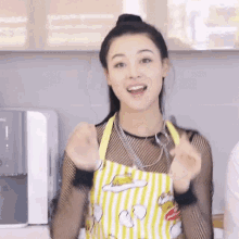 a woman wearing a yellow and white apron is making a heart shape with her hands in a kitchen .