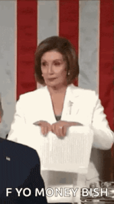 a woman in a white suit is holding a piece of paper in front of a man and says `` f yo money ish '' .