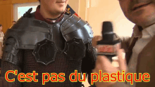 a man in armor is being interviewed by a man in a suit with the words " c'est pas du plastique " in yellow