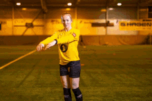 a woman in a yellow jersey stands on a soccer field with a sign in the background that says hagen