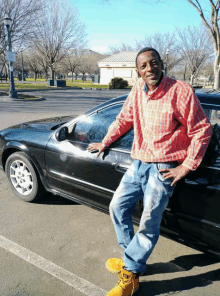 a man in a plaid shirt leans against a car in a parking lot