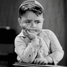 a black and white photo of a young boy sitting at a desk with his hand on his chin .