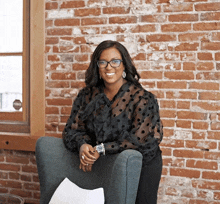 a woman wearing glasses and a black polka dot shirt is sitting in a chair in front of a brick wall