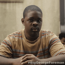 a young man in a striped shirt is sitting with his hands folded in front of a sign that says #biggeorgeforeman