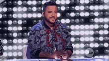 a man with a beard is sitting at a table in front of a stage with lights behind him .