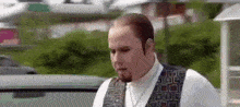 a man with a beard and a vest is standing in front of a car in a parking lot .