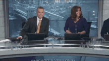 a man and a woman sit at a desk with laptops in front of a chicago 's very own sign