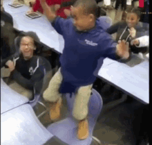 a boy in a blue shirt is jumping in the air while sitting on a chair in a classroom .