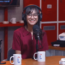 a woman wearing headphones and glasses stands in front of a microphone and two coffee mugs with letters c on them