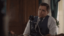 a man in a vest and tie is sitting at a desk talking on an old fashioned telephone