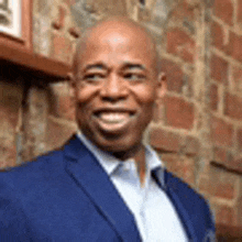 a bald man in a blue suit and white shirt is smiling in front of a brick wall .