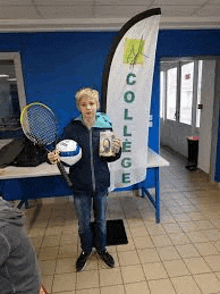 a young boy is holding a tennis racket , a volleyball , and a book .