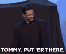 a man in a priest 's robe is standing on a stage and saying `` tommy put 'er there ''