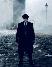 a man in a suit and hat stands on a cobblestone street in the fog