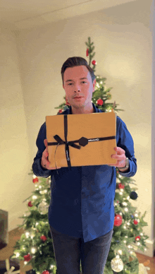 a man in front of a christmas tree holds a gift box with a black bow