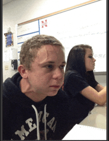 a boy wearing a black shirt with the word mexican on it sits in front of a white board