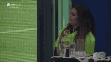 a woman in a green shirt is sitting at a table in front of a screen that says playelli 's