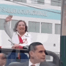 a woman in a white coat is waving from the roof of a car in front of a building that says salud ocupacional .