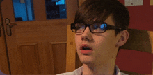 a young boy wearing glasses looks surprised in front of a wooden door