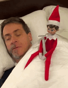 an elf on the shelf is holding a razor next to a sleeping man