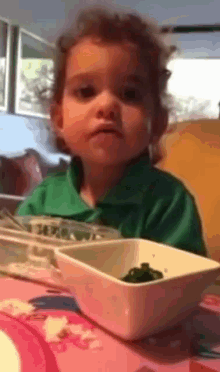 a young boy in a green shirt sits at a table with a bowl of food in front of him