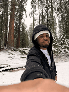 a man wearing a hooded jacket and a white hat takes a selfie in the snow