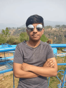 a young man wearing sunglasses stands with his arms crossed in front of a blue railing