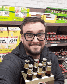 a man in glasses holds a six pack of beer in front of boxes of corn flakes