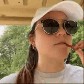 a woman wearing sunglasses , a hat and a white shirt is eating a candy .