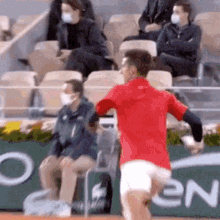 a man in a red shirt is running on a tennis court in front of a crowd wearing masks .