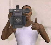 a man in a white tank top is holding a video camera in front of his face and giving a thumbs up