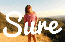 a woman in a pink shirt stands on a hill with the word sure behind her