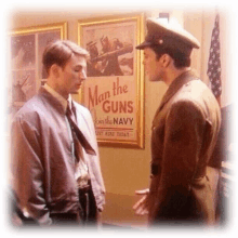 two men talking in front of a poster that says man the guns join the navy