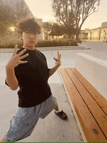 a young man in a black shirt is standing next to a wooden bench and giving a peace sign