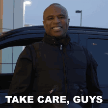 a man in a black vest stands in front of a car with the words " take care guys " written below him