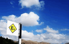 a yellow and white sign with the letter e on it is on a pole in front of a blue sky with clouds .