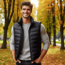 a man wearing a black vest and a grey sweater stands in a park with his hands in his pockets