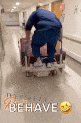 a man is pushing a hospital bed down a hallway with the words they see me behave behind him