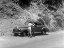 a black and white photo of a woman standing next to a car on the side of a road .