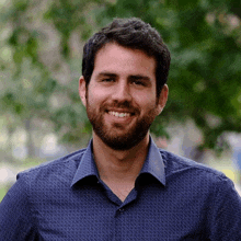 a man with a beard wearing a blue shirt smiles at the camera