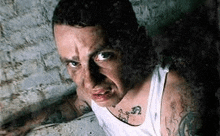 a man with tattoos on his arms and chest is making a funny face while standing in front of a brick wall .