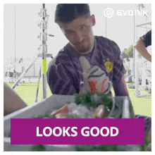 a man in a purple shirt is sitting in front of a container of food and a sign that says looks good .