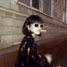 a woman wearing sunglasses smoking a cigarette in front of a brick wall .