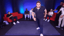 a man in a black shirt is dancing in front of a group of men sitting on red couches