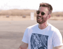 a man with a beard wearing sunglasses and a white t-shirt is smiling .