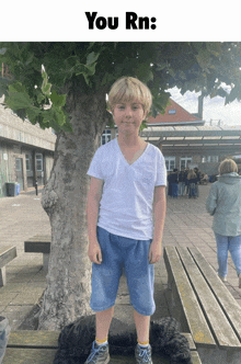 a boy in a white shirt and blue shorts stands in front of a tree with the words you rn below him