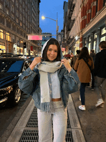 a woman wearing a scarf and a denim jacket stands on a street