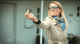 a woman wearing glasses and a trench coat shows her middle finger