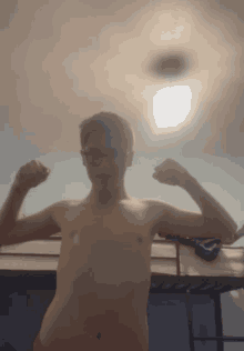 a shirtless man with glasses flexes his muscles in front of a light